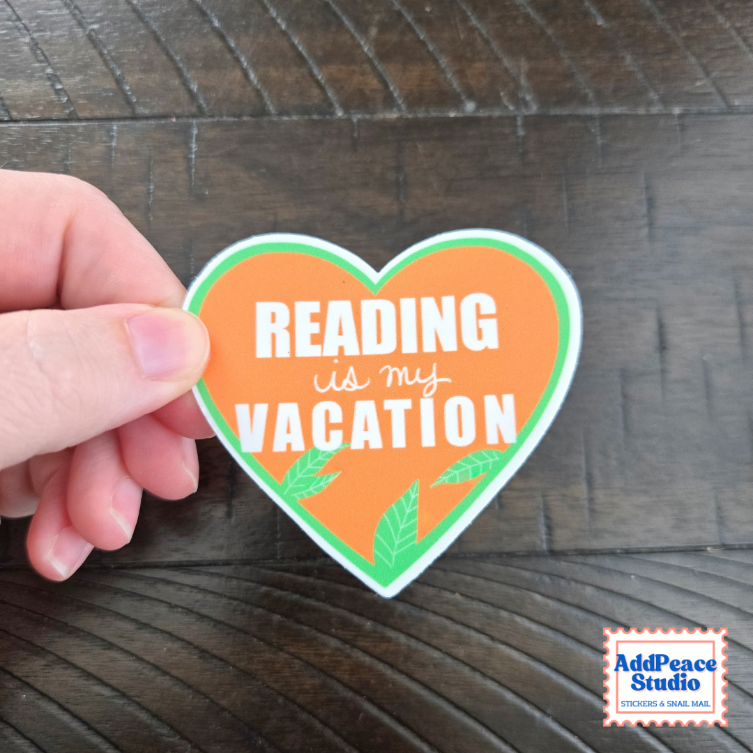 A hand holds a heart-shaped sticker with the text 'READING is my VACATION' in white on an orange background with green leaf accents, set against a dark wood surface. AddPeace Studio logo is visible in the bottom right corner.