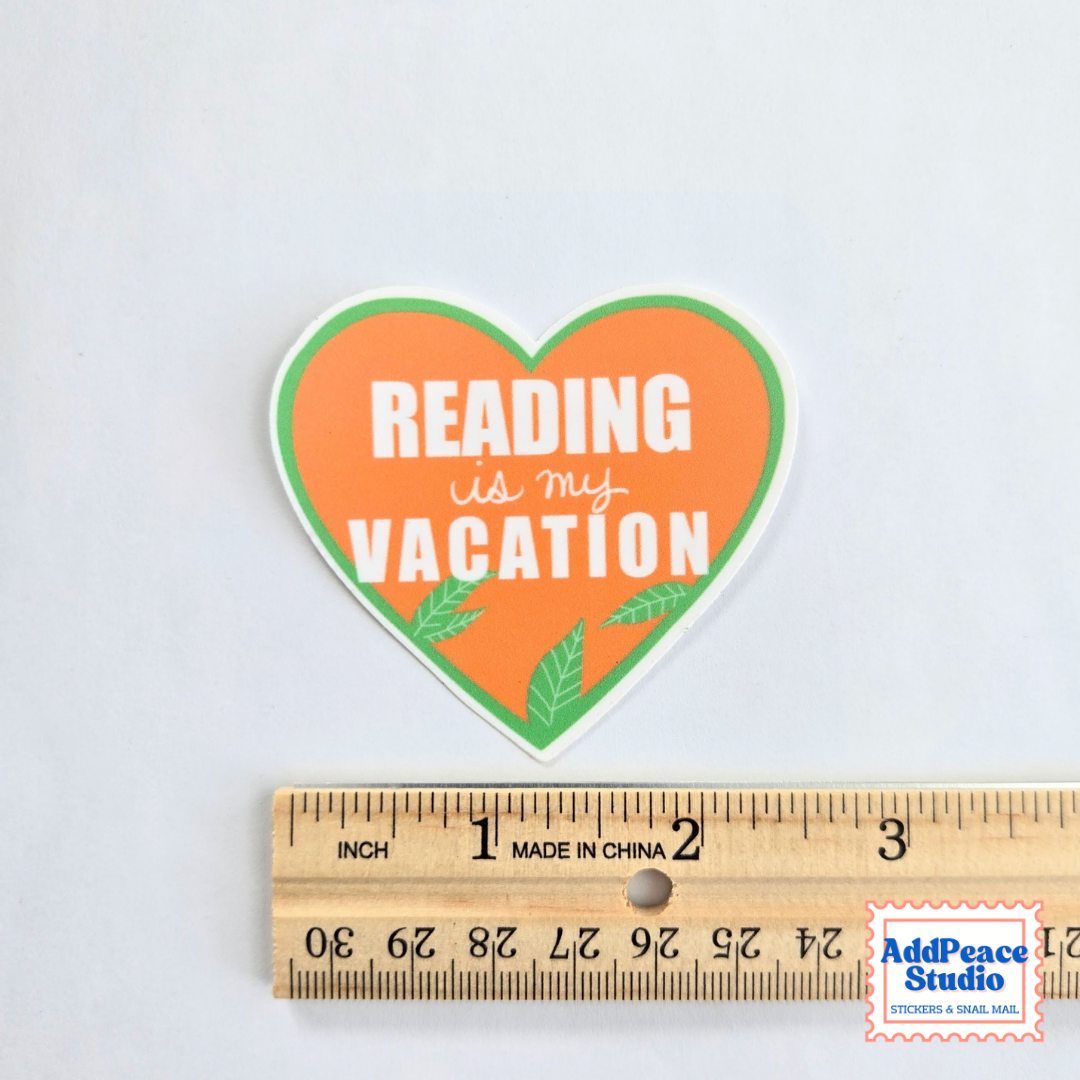 A heart-shaped sticker with the text 'READING is my VACATION' in white lettering on an orange background with green leaf accents, shown above a wooden ruler for scale on a light gray background.