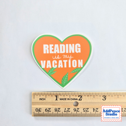 A heart-shaped sticker with the text 'READING is my VACATION' in white lettering on an orange background with green leaf accents, shown above a wooden ruler for scale on a light gray background.