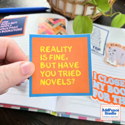 A hand holds a square orange sticker with a blue border and yellow text that reads, 'REALITY IS FINE, BUT HAVE YOU TRIED NOVELS?' in front of an open notebook with colorful stickers.