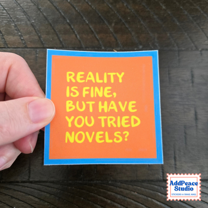 A hand holds a square sticker with an orange background, blue border, and yellow text that reads 'REALITY IS FINE, BUT HAVE YOU TRIED NOVELS?' on a dark wood surface. An AddPeace Studio logo is visible in the bottom right corner.
