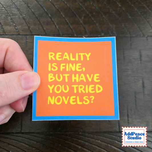 A hand holds a square sticker with an orange background, blue border, and yellow text that reads 'REALITY IS FINE, BUT HAVE YOU TRIED NOVELS?' on a dark wood surface. An AddPeace Studio logo is visible in the bottom right corner.
