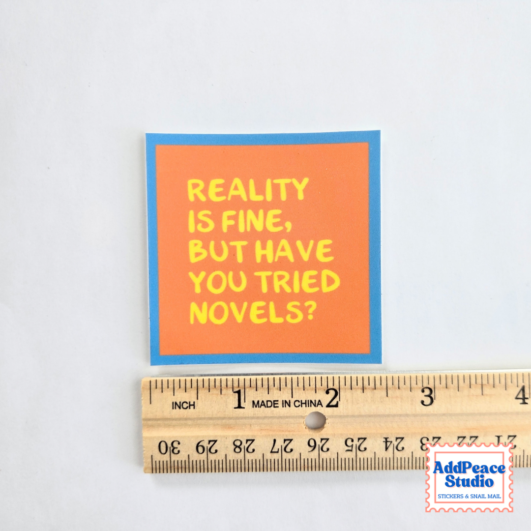 A square sticker with the text 'REALITY IS FINE, BUT HAVE YOU TRIED NOVELS?' in yellow on an orange background with a blue border, shown above a wooden ruler for scale on a white background.