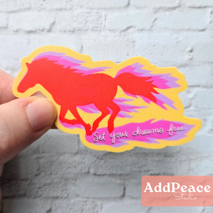Hand holding a colorful horse-shaped sticker with 'Set your dreams free' text against a white brick wall.