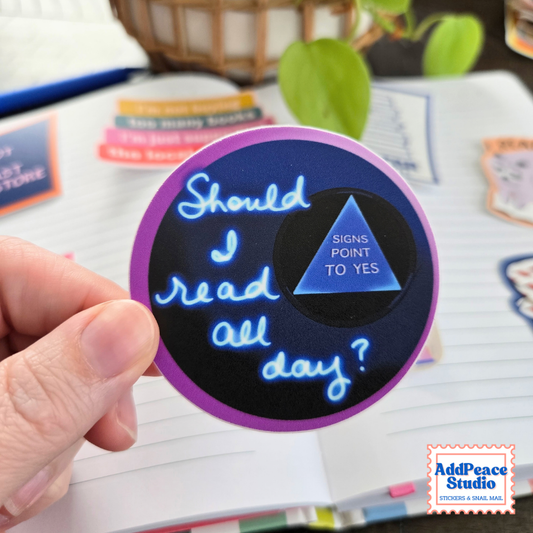 A hand holds a round sticker with a purple border and a black center. The sticker features a blue triangle with the text 'SIGNS POINT TO YES' and blue cursive text reading 'Should I read all day?'.