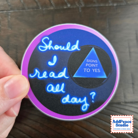 A hand holds a round sticker with a dark blue background, purple border, and neon-style blue text reading 'Should I read all day?'. A blue triangle in the center says 'SIGNS POINT TO YES'.