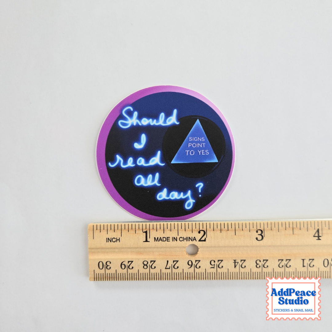 A round sticker with a purple border and a black center, featuring the text 'Should I read all day?' in blue script and a blue triangle that says 'SIGNS POINT TO YES.' A wooden ruler is placed below it to show scale, and a small 'AddPeace Studio' logo appears in the bottom right corner.