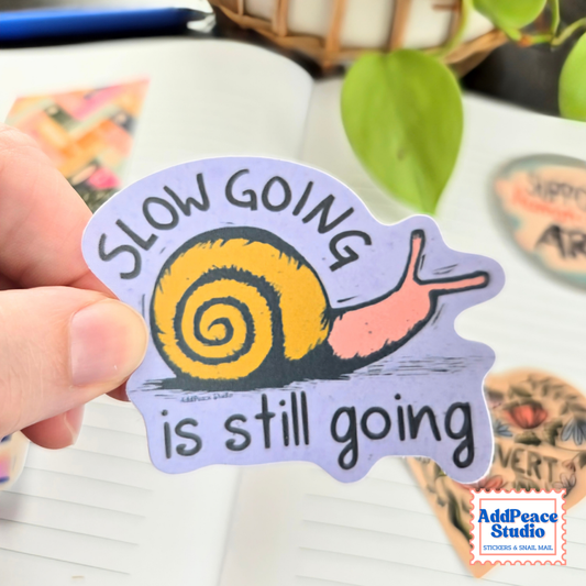 A hand holds a die-cut sticker featuring a snail with a yellow shell and pink body on a lavender background, with the text 'SLOW GOING is still going'. The scene is set on a desk with an open notebook, green leaves, and other stickers in the background.