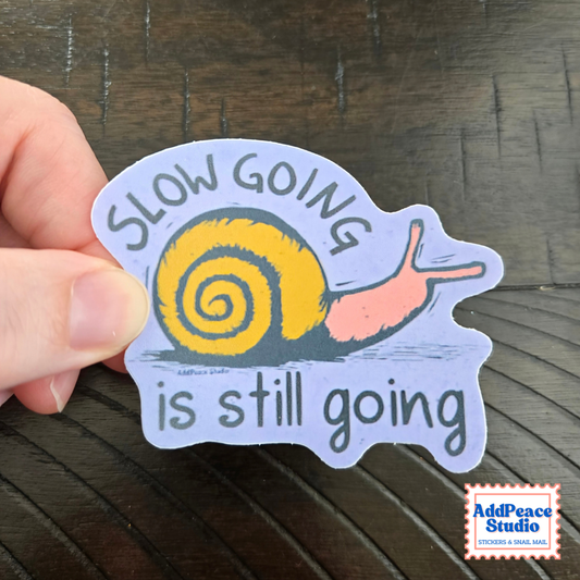 A hand holds a die-cut sticker featuring a snail with a yellow shell and pink body on a light lavender background, with the text 'SLOW GOING is still going'. The sticker is photographed over a dark wood surface, and an 'AddPeace Studio' logo is visible in the bottom right corner.