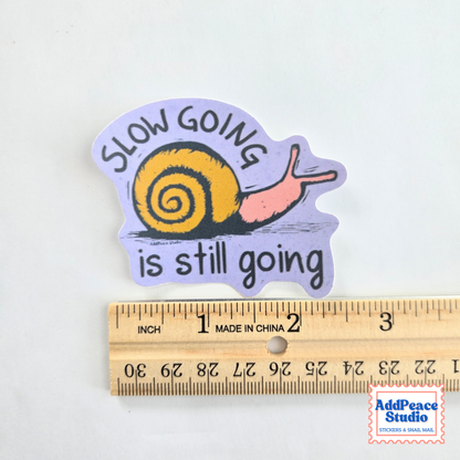 A die-cut sticker featuring a yellow snail on a light purple background with the text 'SLOW GOING is still going' in black. The sticker is shown above a wooden ruler to indicate its size, on a plain white background.