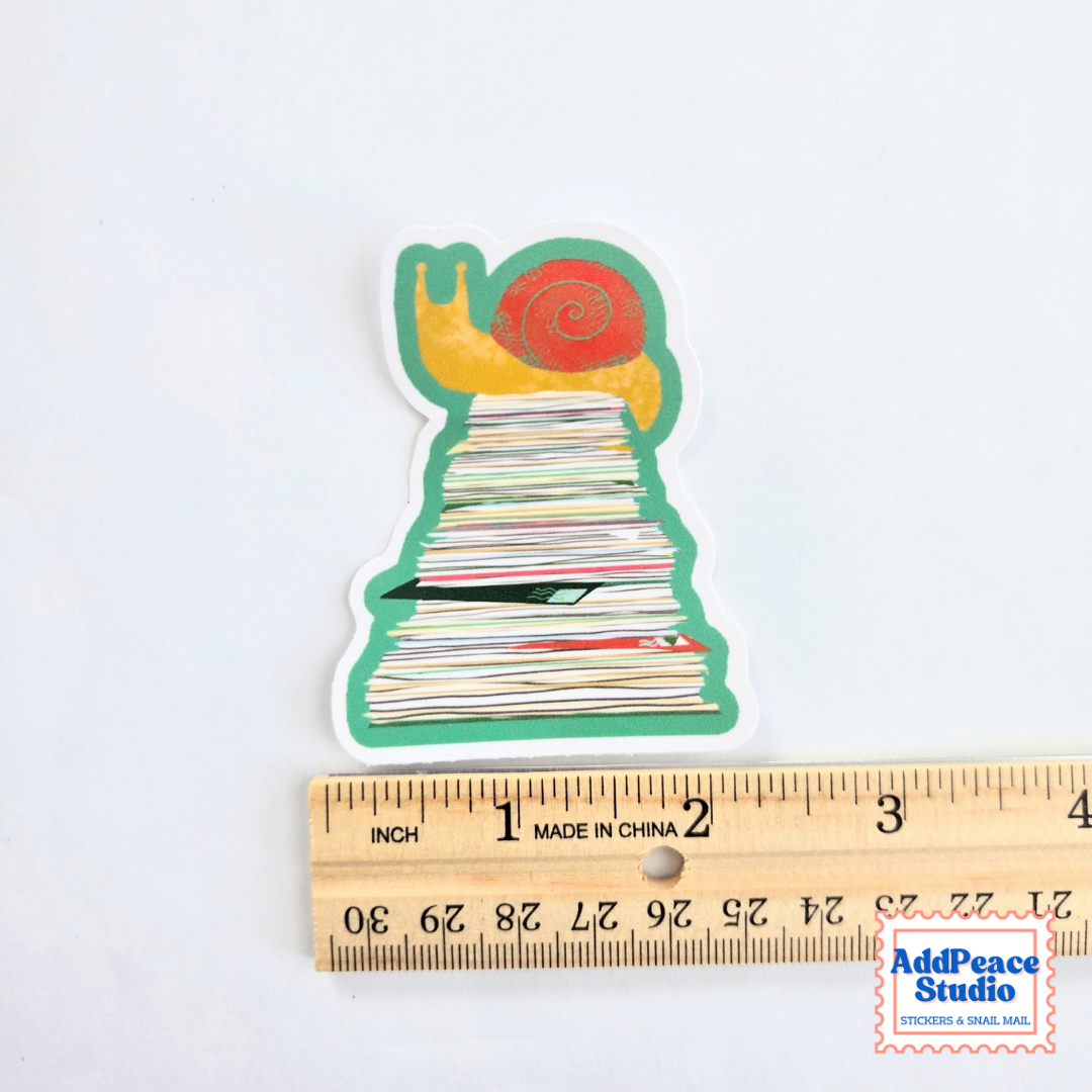 A die-cut sticker shaped like a snail on top of a stack of letters, shown next to a wooden ruler for scale on a white background.