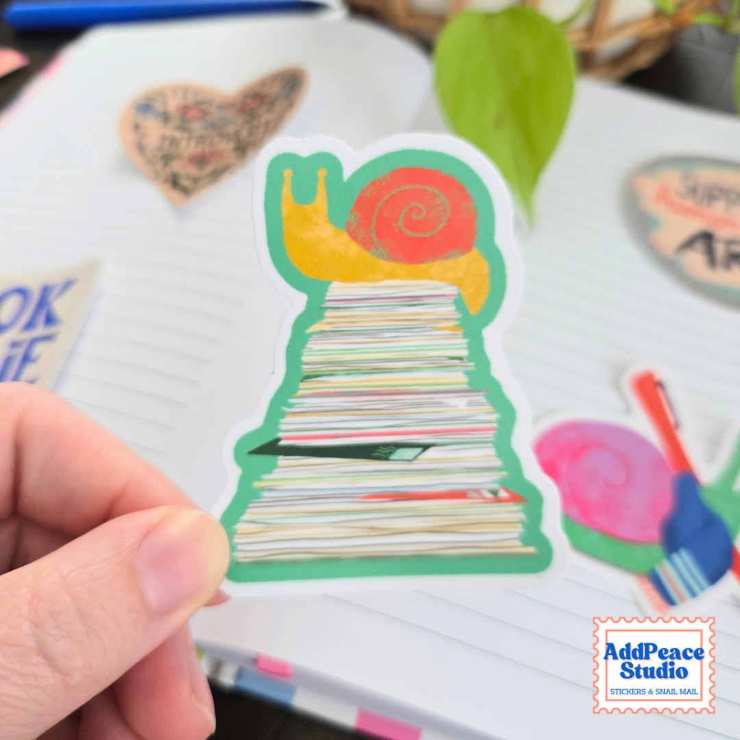 A hand holds a die-cut sticker shaped like a snail on top of a stack of mail with a lined notebook and other colorful stickers in the background. The AddPeace Studio logo is visible in the bottom right corner.
