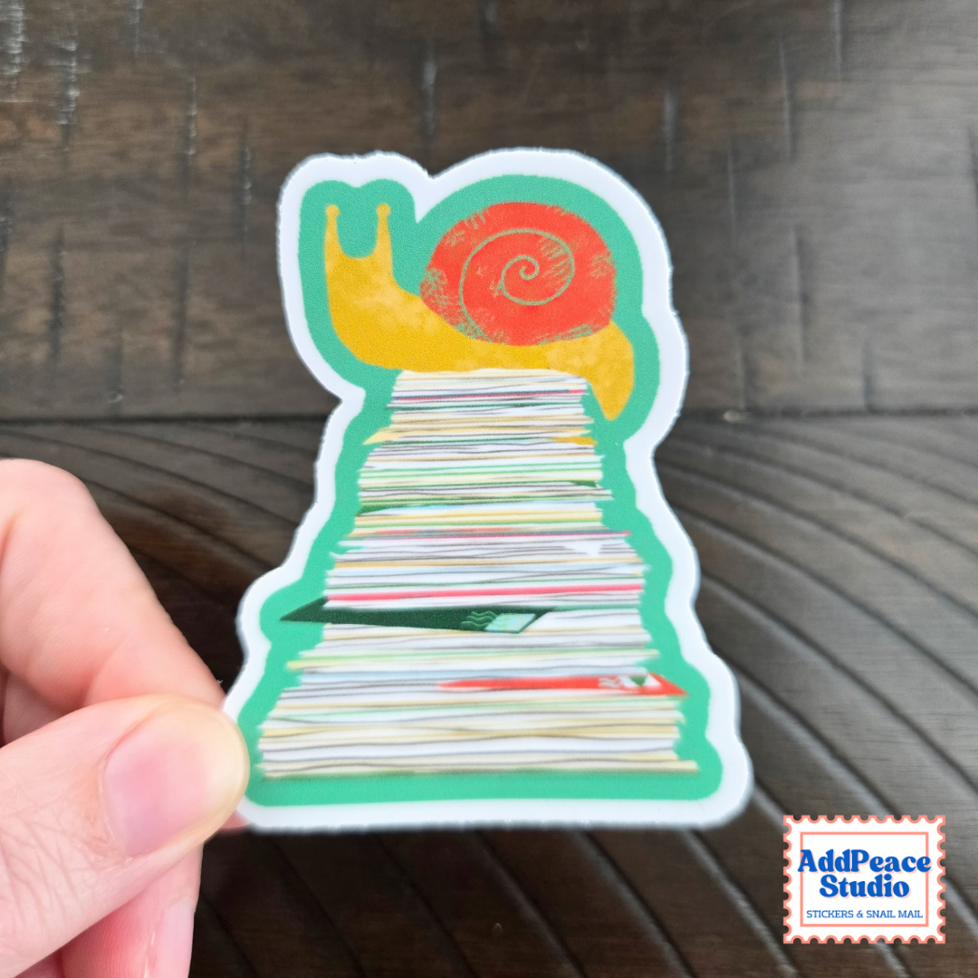 A hand holds a die-cut sticker shaped like a snail on top of a stack of mail, with a red shell and yellow body, against a dark wood background. An AddPeace Studio logo is visible in the bottom right corner.
