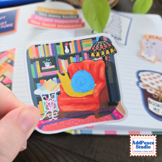 A hand holds a die-cut sticker featuring a cozy book-themed illustration with a red armchair, bookshelves, and a snail reading mail. The sticker has a white border and the AddPeace Studio logo is visible in the bottom right corner.