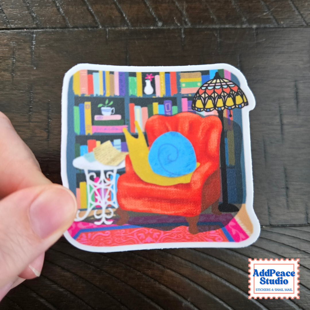 A hand holds a die-cut sticker featuring a colorful cozy library or reading nook illustration with a red armchair, bookshelves, lamp, and patterned rug with a snail opening its mail. The sticker is shown against a dark wood background, with an AddPeace Studio logo in the bottom right corner.