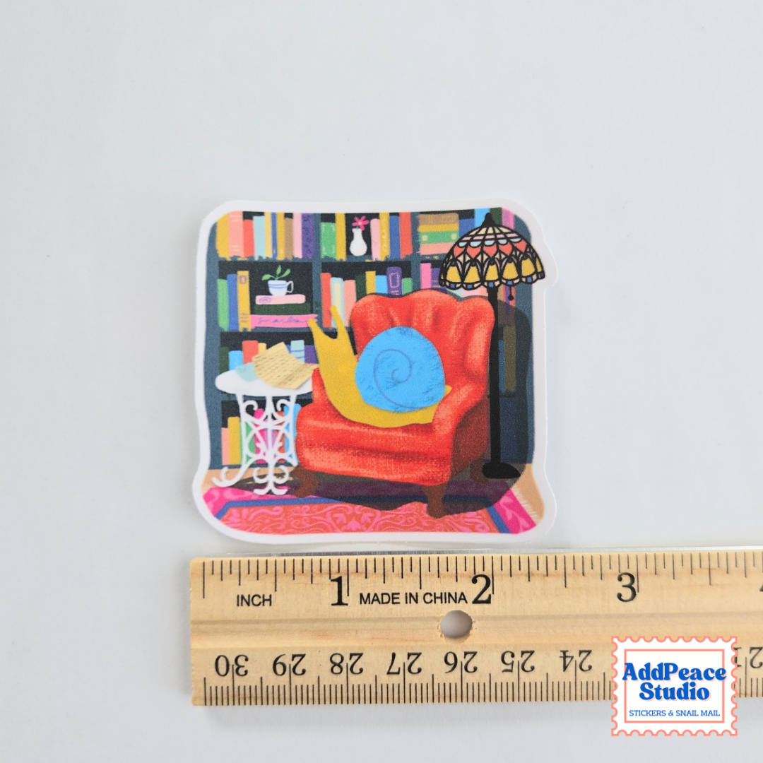 A die-cut sticker featuring a colorful illustration of a cozy living room scene with a red armchair, bookshelves, and a lamp, with a snail reading mail, shown above a wooden ruler for scale on a light gray background.