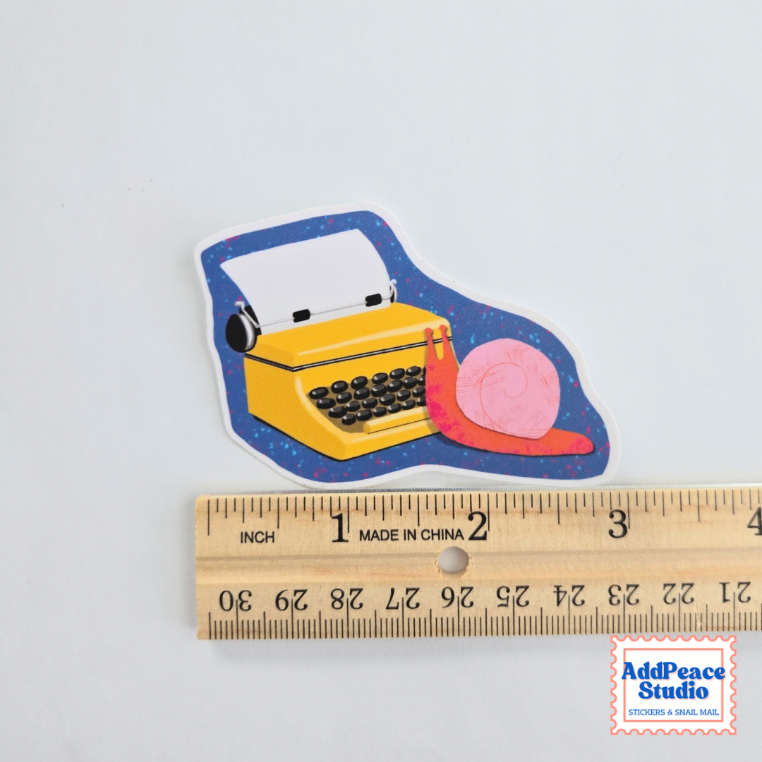 A die-cut sticker featuring a yellow typewriter and a pink snail on a blue speckled background, shown above a wooden ruler for scale. The AddPeace Studio logo is in the bottom right corner.