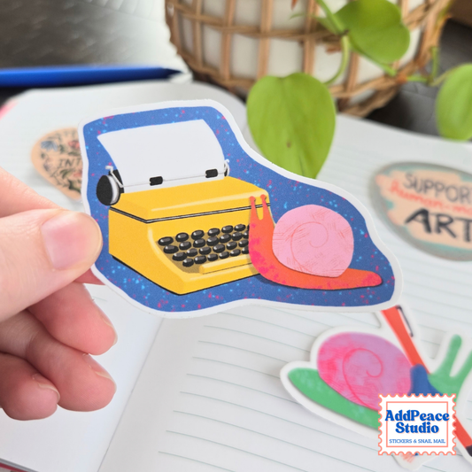 A hand holds a die-cut sticker featuring a yellow typewriter and a pink snail on a blue background, with other stickers and a notebook visible in a styled desk setting. The AddPeace Studio logo appears in the bottom right corner.