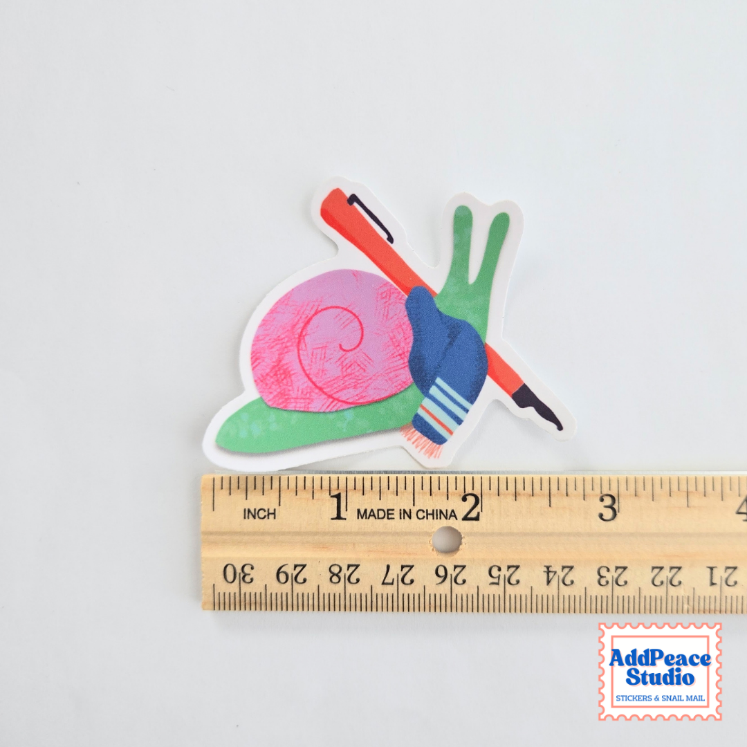 A colorful snail-shaped sticker shown next to a wooden ruler to indicate scale, on a plain white background. The sticker features a pink snail shell, green body, and a red fountain pen.
