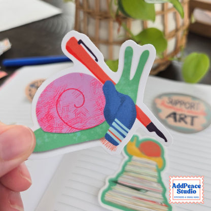 A hand holds a colorful snail-shaped sticker featuring a pink shell, green body, and red fountain pen, with other stationery items blurred in the background.