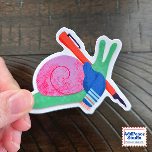 A hand holds a die-cut sticker featuring a colorful snail design with a pink shell, green body, holding a red fountain pen, set against a dark wood background. An 'AddPeace Studio' logo is visible in the bottom right corner.