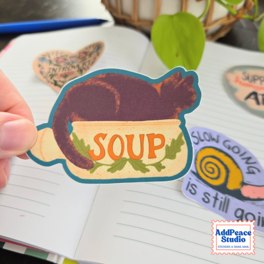 A hand holds a die-cut sticker shaped like a soup bowl with a dark cat sitting inside and the word 'SOUP' in orange letters. Other stickers are scattered on an open notebook in the background, with a plant leaf and basket visible.