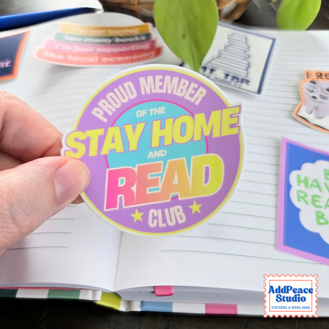 A hand holds a colorful round sticker that reads 'PROUD MEMBER OF THE STAY HOME AND READ CLUB' over an open notebook with other stickers visible in the background.