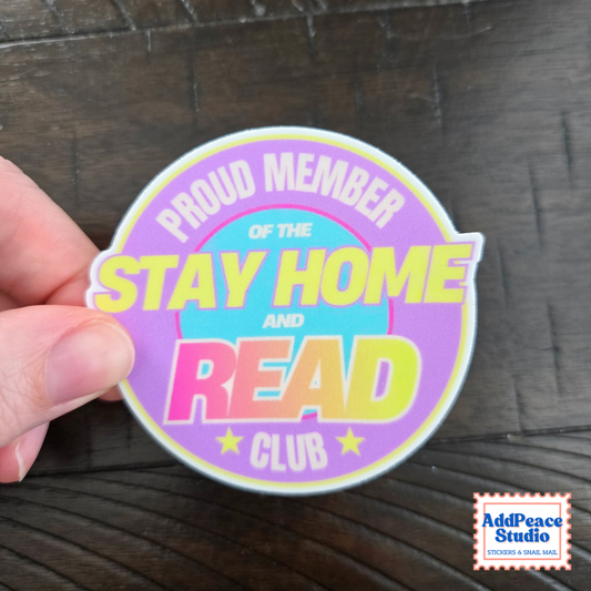 A hand holds a die-cut sticker that reads 'PROUD MEMBER OF THE STAY HOME AND READ CLUB' in pastel colors on a dark wood background. A small 'AddPeace Studio' logo appears in the bottom right corner.