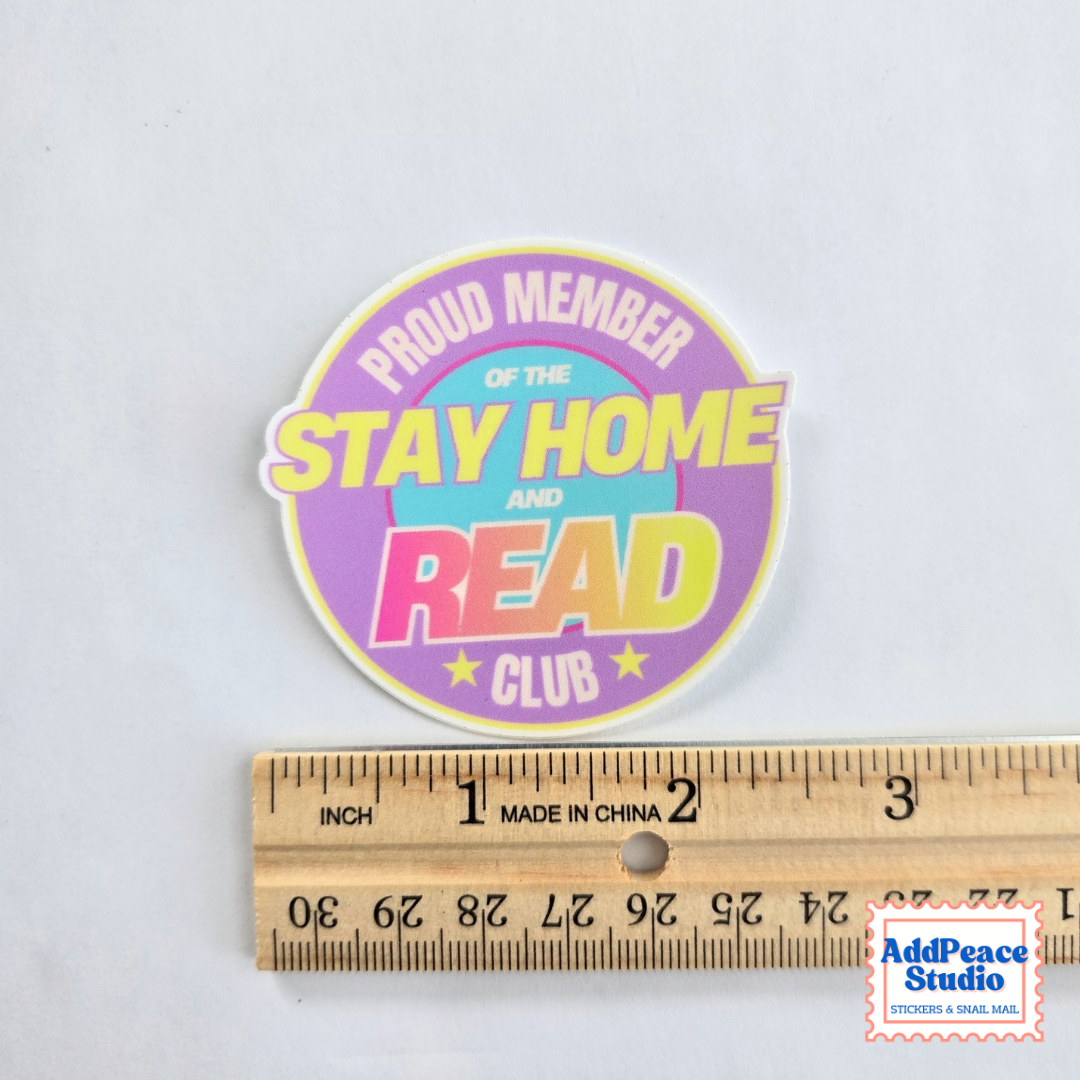 A round die-cut sticker reading 'PROUD MEMBER OF THE STAY HOME AND READ CLUB' in pastel colors, shown above a wooden ruler for scale on a white background.