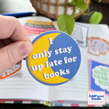 A hand holds a round sticker that reads 'I only stay up late for books' in white text over a yellow and blue crescent design. The background shows an open notebook with other colorful stickers and a blurred plant.