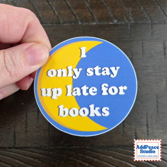 A hand holds a round sticker with a blue and yellow crescent moon design and the text 'I only stay up late for books' in white lettering, set against a dark wood background. An 'AddPeace Studio' logo is visible in the bottom right corner.