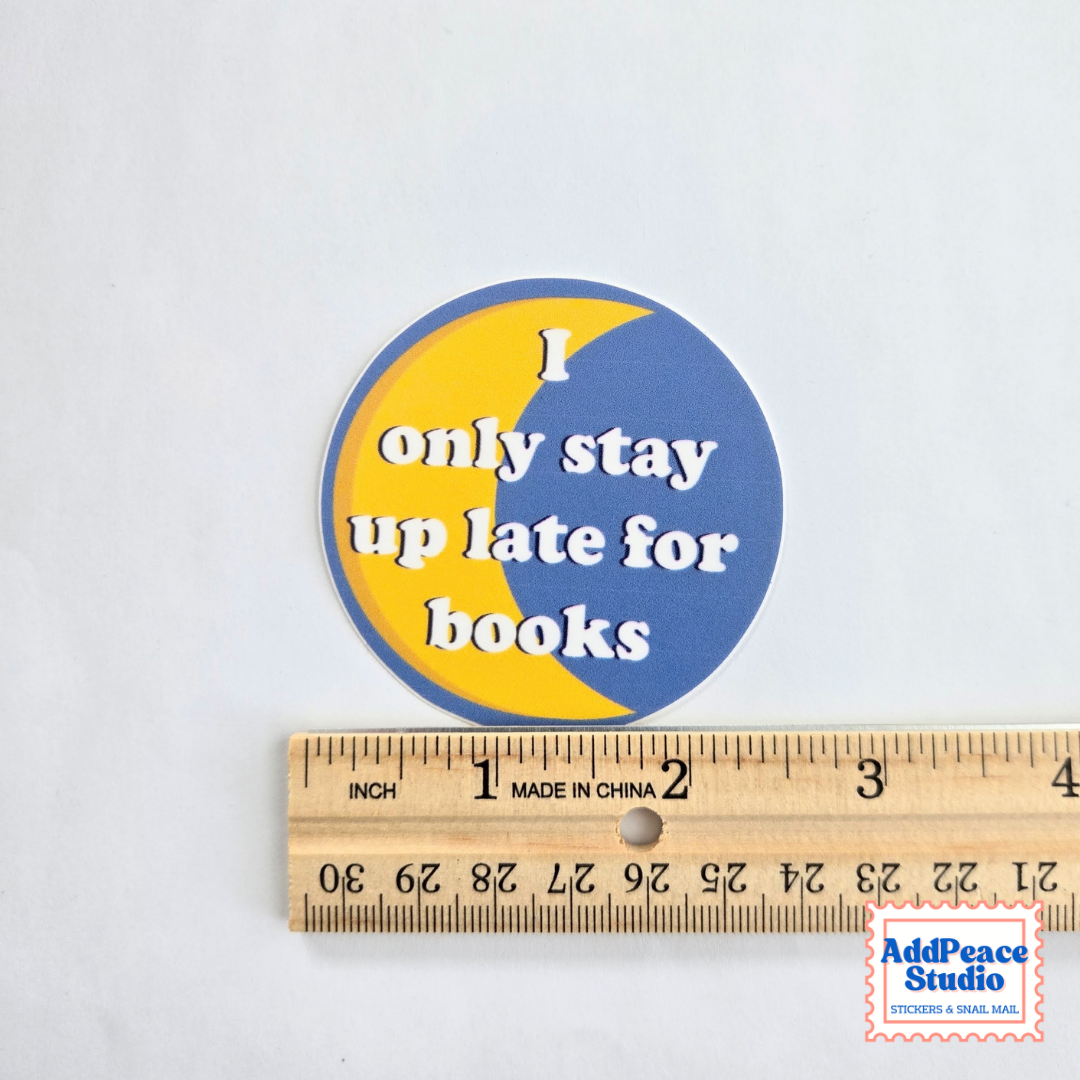 A round sticker with a blue and yellow crescent moon design and the text 'I only stay up late for books' in white, shown above a wooden ruler for scale on a white background.