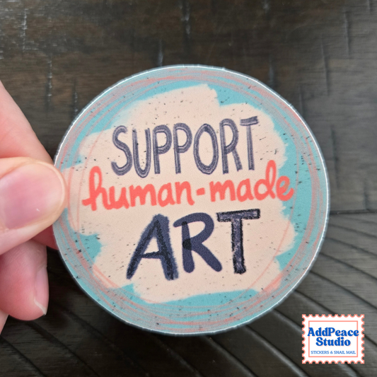A hand holds a round sticker that reads 'SUPPORT human-made ART' in stylized lettering. The sticker has a light pink background with blue and coral accents, set against a dark wood surface.
