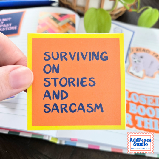 A hand holds an orange square sticker with a yellow background with dark blue text that says Surviving on stories and sarcasm. The background is an open notebook with colorful stickers and plans.
