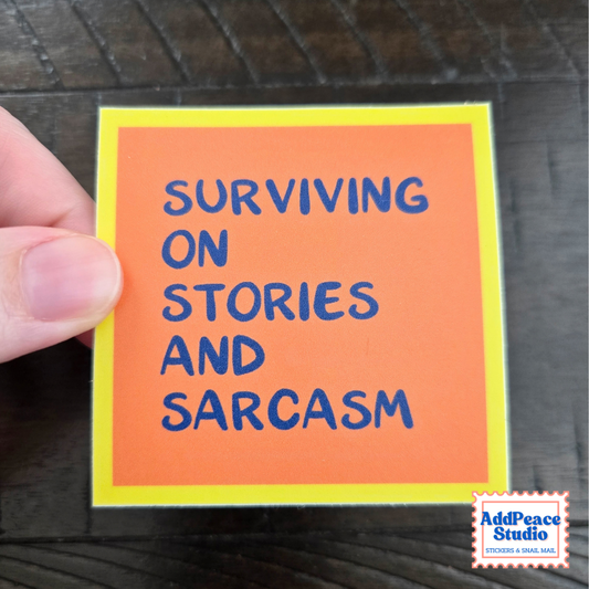 A hand holds a square orange sticker with a yellow border and blue handwritten text reading 'SURVIVING ON STORIES AND SARCASM' against a dark wood background. An 'AddPeace Studio' logo appears in the bottom right corner.