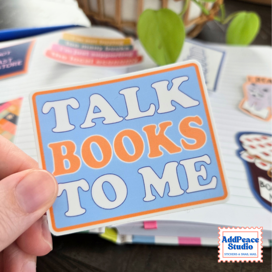 A hand holds a rectangular sticker that reads 'TALK BOOKS TO ME' in white and orange lettering on a light blue background with an orange border. The scene is a styled desk setup with notebooks, pens, and a plant in the background.