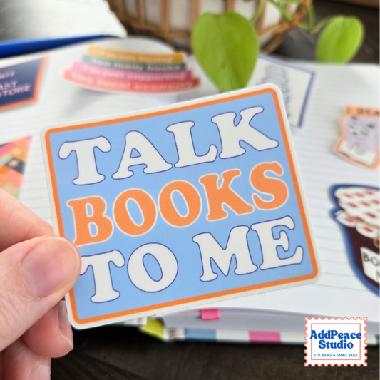 A hand holds a rectangular sticker that reads 'TALK BOOKS TO ME' in white and orange lettering on a light blue background with an orange border. The scene is a styled desk setup with notebooks, pens, and a plant in the background.