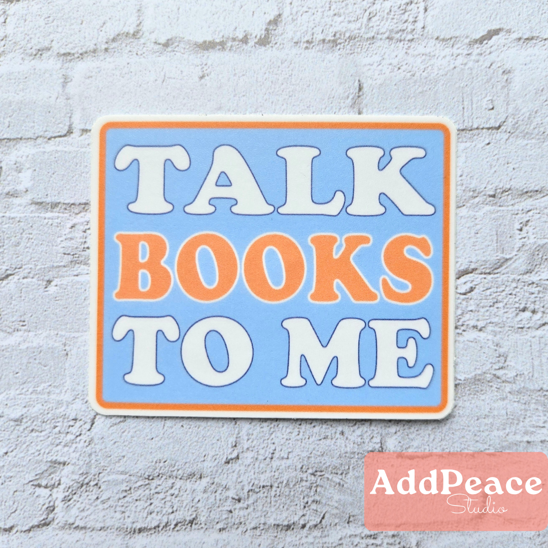 Decorative sign with 'Talk Books to Me' text on a light blue background, placed on a textured white surface.