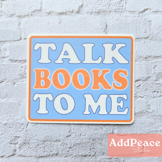 Decorative sign with 'Talk Books to Me' text on a light blue background, placed on a textured white surface.