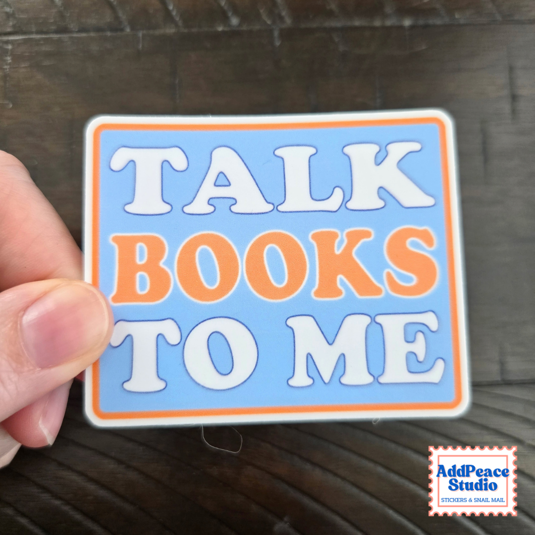 A hand holds a rectangular sticker with the text 'TALK BOOKS TO ME' in white and orange lettering on a light blue background with an orange border. The sticker is photographed over a dark wood surface, with an 'AddPeace Studio' watermark in the bottom right corner.