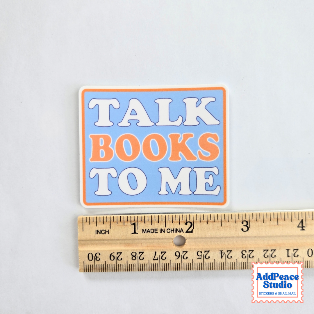 A square sticker with the text 'TALK BOOKS TO ME' in white and orange lettering on a light blue background with an orange border, shown next to a wooden ruler for scale on a white background.