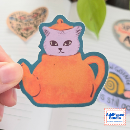 A hand holds a die-cut sticker featuring a purple cat sitting inside an orange teapot, outlined in dark teal. The sticker is shown against a white lined notebook background with other stickers blurred in the background.