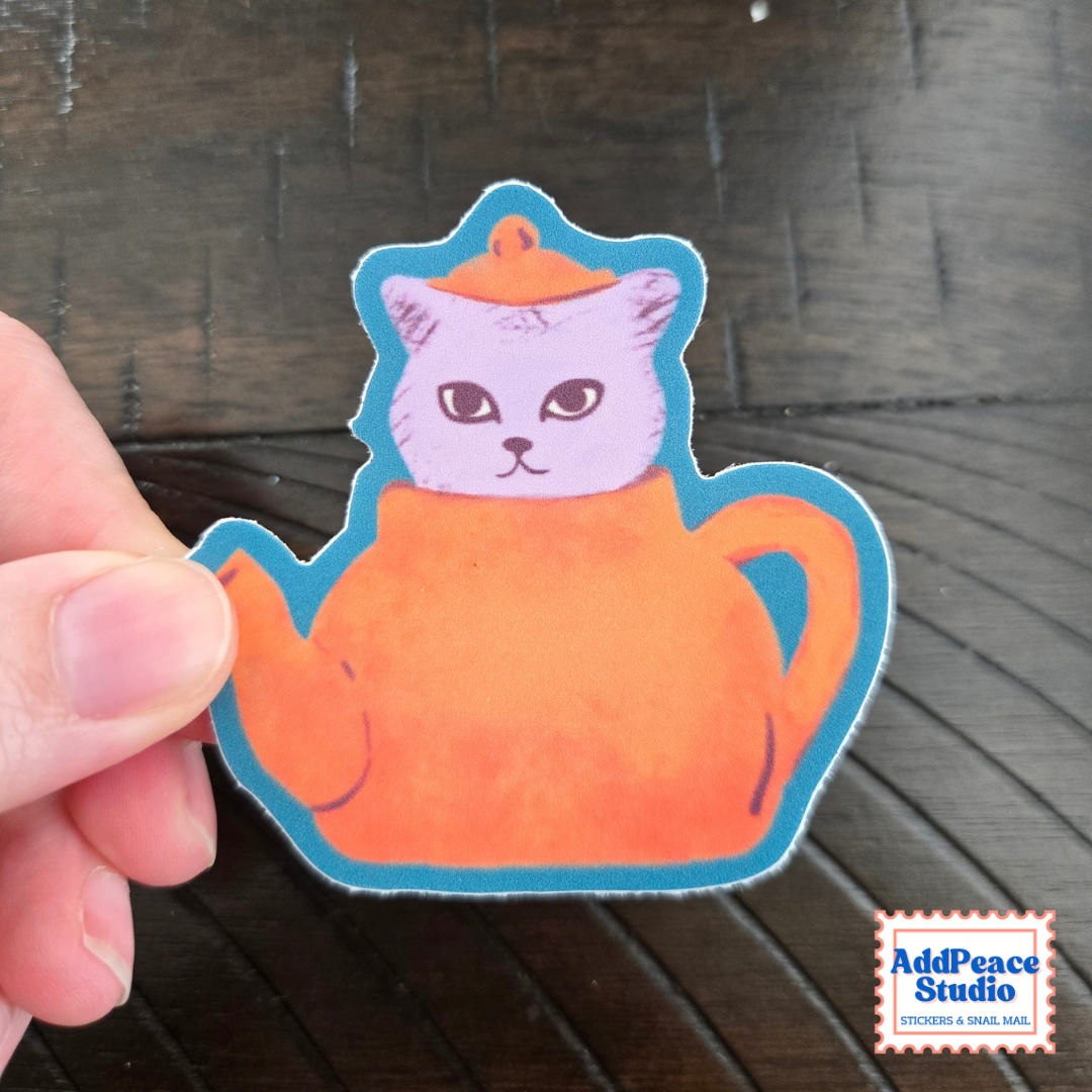 A hand holds a die-cut sticker featuring a white cat with large eyes inside an orange teapot, outlined in blue, against a dark wood background. An AddPeace Studio logo is visible in the bottom right corner.