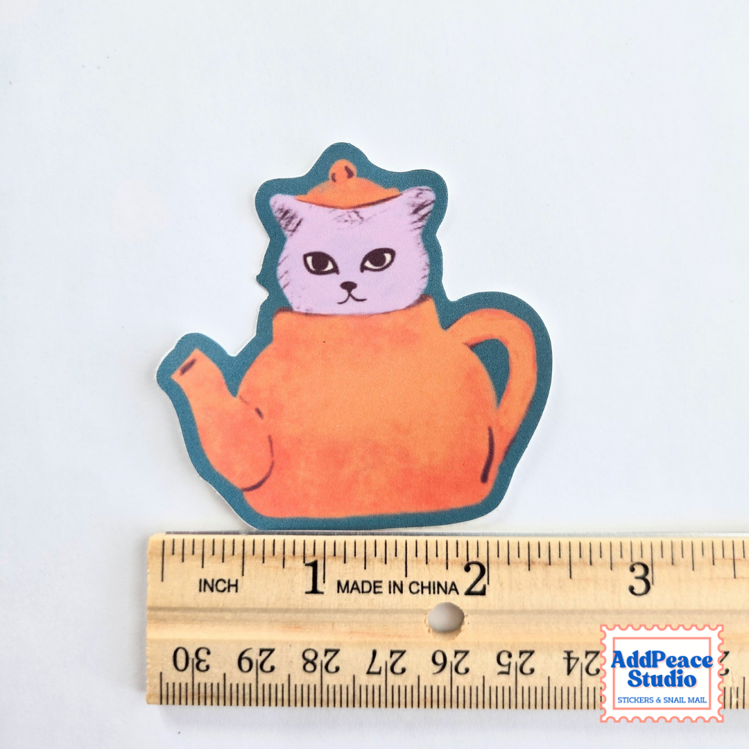 A small die-cut sticker or decal featuring a cartoon cat inside an orange teapot, shown above a wooden ruler for scale on a white background. The sticker has a dark teal outline and a light lavender/pink cat face.