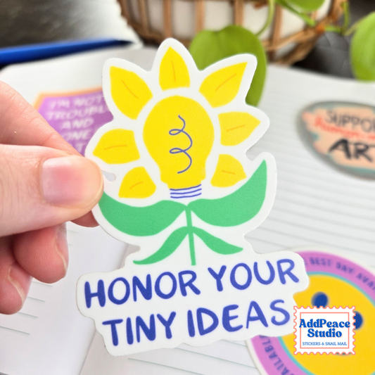 A hand holds a die-cut sticker shaped like a flower with a lightbulb in the center and the text 'HONOR YOUR TINY IDEAS' in blue. The sticker has yellow petals, green leaves, and a white border.