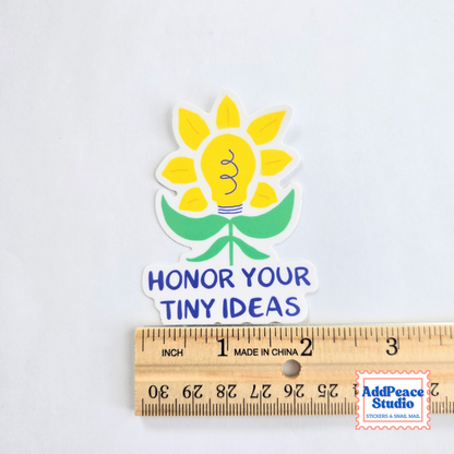 A die-cut sticker featuring a yellow sunflower with a lightbulb in the center, green leaves, and the text 'HONOR YOUR TINY IDEAS' in blue. The sticker is displayed above a wooden ruler to show its size, on a plain white background.