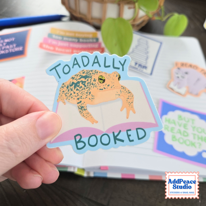 A hand holds a die-cut sticker featuring a toad sitting on an open book with the words 'TOADALLY BOOKED' in dark teal lettering. The sticker is shown in front of a decorated notebook with other colorful stickers, and a plant is blurred in the background.