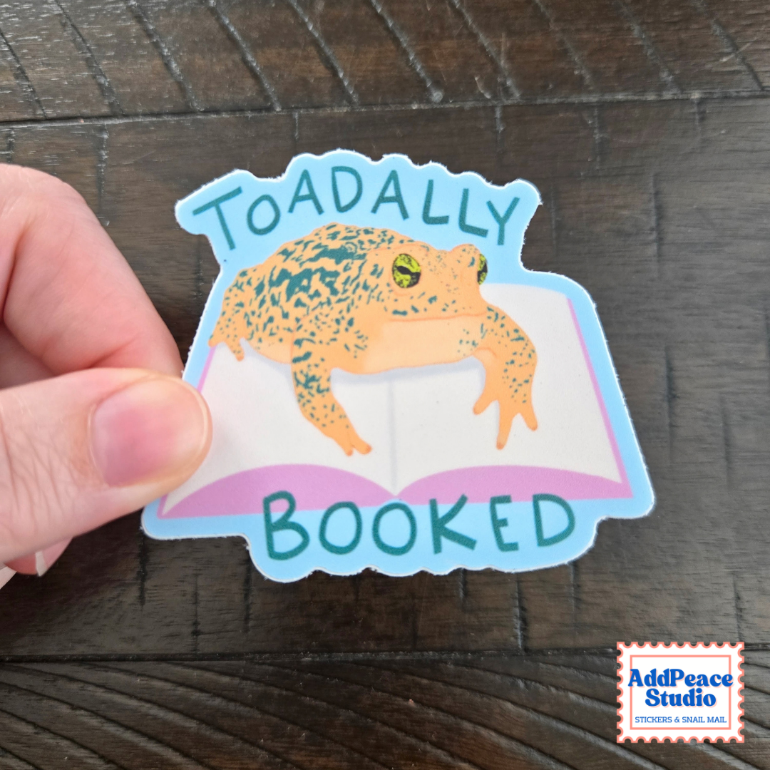 A hand holds a die-cut sticker featuring a toad sitting on an open book with the words 'TOADALLY BOOKED' in teal, on a dark wood background. A small 'AddPeace Studio' logo is visible in the bottom right corner.