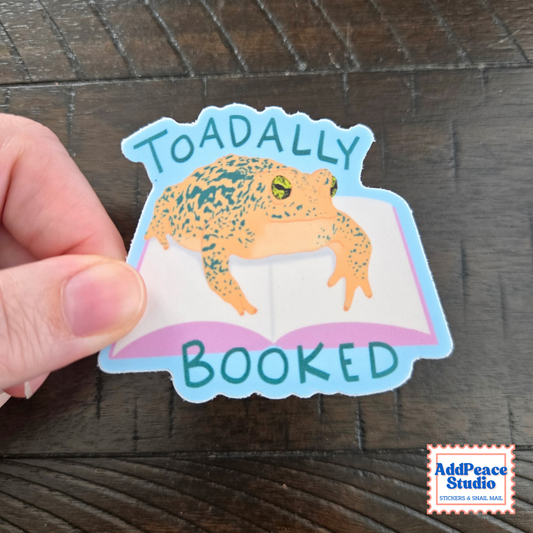 A hand holds a die-cut sticker featuring a toad sitting on an open book with the words 'TOADALLY BOOKED' in teal, on a dark wood background. A small 'AddPeace Studio' logo is visible in the bottom right corner.