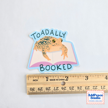 A die-cut sticker featuring a toad sitting on an open book with the words 'TOADALLY BOOKED' in dark teal lettering, shown above a wooden ruler for scale on a white background.